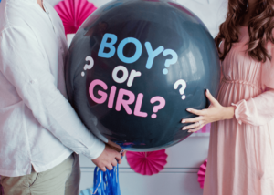 A person in a white shirt and another in a pink dress hold a large black balloon with the text "BOY? or GIRL?" in blue and pink letters. The background, featuring pink decorations attached to a white surface, adds a delightful touch to this fun gender reveal idea.