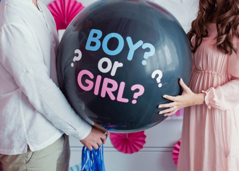 A person in a white shirt and another in a pink dress hold a large black balloon with the text "BOY? or GIRL?" in blue and pink letters. The background, featuring pink decorations attached to a white surface, adds a delightful touch to this fun gender reveal idea.