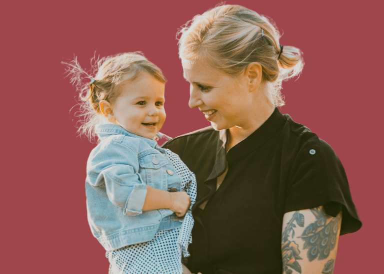 A woman with a tattoo on her arm is holding a small child. The woman is looking at the child and smiling, while the child, wearing a denim jacket and a polka dot outfit, looks to the side. The background is a solid reddish color, evoking themes of maternal mental health through serene connection.