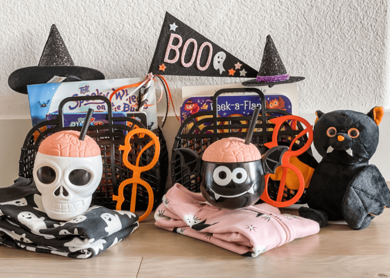 A Halloween-themed setup featuring two black baskets with spooky books and decorations. In front are two cups shaped like a skull and a bat, Halloween-themed glasses, a plush black cat, and Halloween-patterned cloths. A "BOO" decoration and witch hats complete the scene, perfect as Halloween gifts for kids.