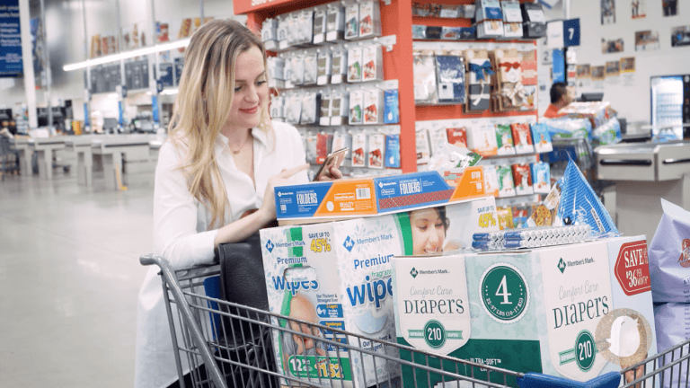 A woman in a white shirt stands beside a shopping cart filled with various baby products like diapers and wipes. She is looking at her phone, presumably checking her to-do list. The background shows the aisles of Sam's Club, complete with shelves and displays, including magazine racks and other customers.
