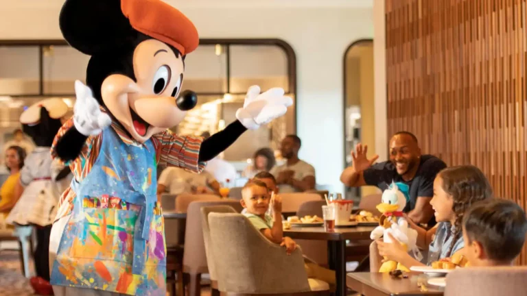 An animated character dressed as an artist mouse interacts with children and families in a restaurant setting. The character wears a colorful outfit and a red beret, resembling a scene from Disney character dining. Several tables of diners watch and wave, creating a lively and cheerful atmosphere.