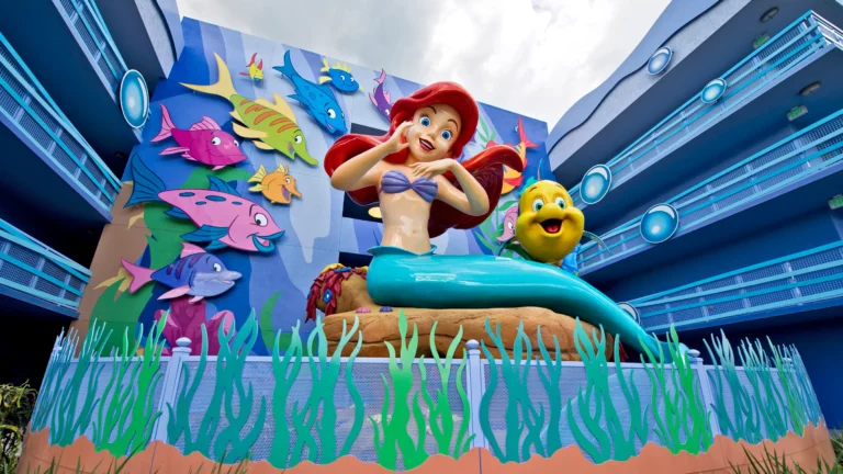A colorful sculpture of Ariel and Flounder from "The Little Mermaid" is displayed in front of an animated mural featuring various fish. Ariel is depicted as a mermaid with red hair and a purple seashell bra, while Flounder smiles next to her, capturing the magic often found at the best Disney World hotels.