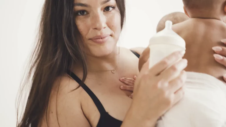 A woman with long dark hair, wearing a black top, is holding an infant who is facing away from the camera. She holds a baby bottle in her other hand and looks into the camera with a serene expression. The background is blurred and light-colored, creating a subtle yet radiant scene reminiscent of celebrity photos.
