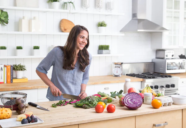A woman, Stephanie Middleberg, with long dark hair is smiling while chopping vegetables in a modern kitchen. The counter is filled with fresh produce, including tomatoes, red cabbage, and leafy greens. Shelves with various kitchen items are visible in the background.