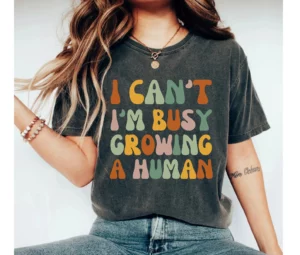 A woman is wearing a dark gray t-shirt with colorful text that reads, I can't, I'm busy growing a human. As one of her pregnancy announcement shirts, she proudly displays her inner forearm tattoo while sporting a necklace and bracelet.