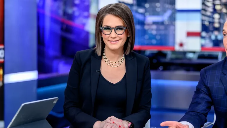 Jessica Tarlov with short brown hair and glasses is sitting in front of a blurred background of news studio screens. They are wearing a black blazer, a black shirt, and a pearl necklace. Additionally, part of someones shoulder and arm can be seen on the right.
